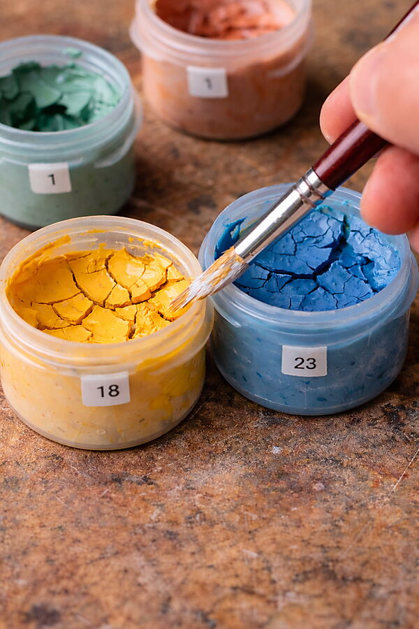 Nahaufnahme von geöffneten Farbtöpfchen mit rissig eingetrockneter Acrylfarbe; ein Pinsel berührt die Oberfläche – typisches Problem bei Malen-nach-Zahlen-Sets.