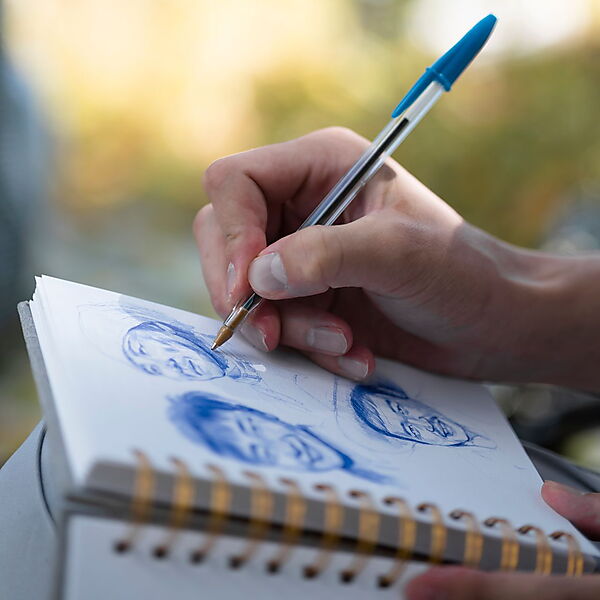 Eine Hand zeichnet mit einem blauen Kugelschreiber mehrere schnelle Porträtskizzen in ein Skizzenbuch.