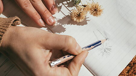 Hände zeichnen mit Bleistift in ein Notizbuch und nutzen einfache natürliche Motive als Vorlage.