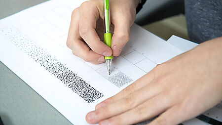 Hand zeichnet mit einem Bleistift Übungslinien auf kariertem Papier und trainiert Druck, Linienführung und Schraffur.