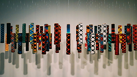 Farbig gemusterte, zylindrische Ornamente hängen in einer Reihe vor einer hellen Wand.